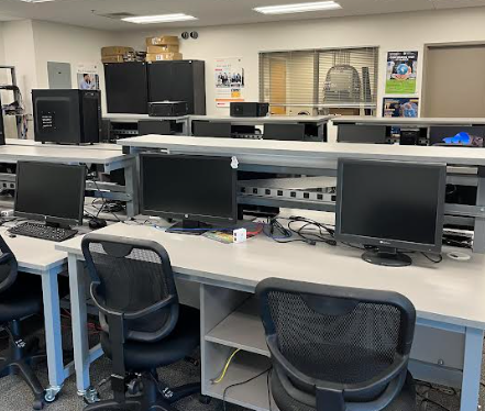 A row of computers in a classroom with roller chairs in front of them.
