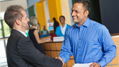 Two men in professional attire shake hands at a job fair. One of the men holds a resume to give to the potential employer.
