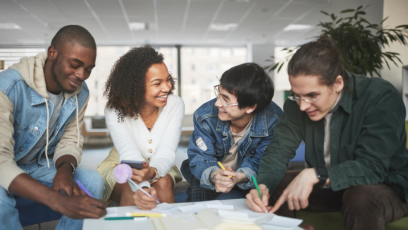 Four young adults sitting together at a table in a bright, modern office space, smiling and collaborating on a creative project with notebooks, papers, and colored pens