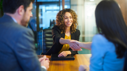 A job candidate sits across from two hiring team members. The candite is smiling and shaking hands with one of the hiring team members.