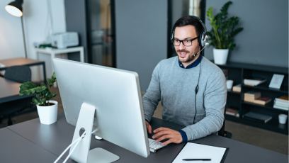 An IT helpdesk worker sits at his desk in from of a computer and talks into the headset he is wearing. There is a clipboard and pen on the desk to his left.