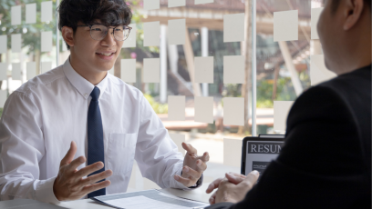 Man in a white shirt, dark tie, and glasses tells a story in an interview and uses his hands for emphasis. The interviewer, who wears a dark suit and is facing the candidate, listens with his hand folded in from of him.