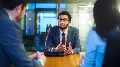 A job candidate in a suit and tie with glasses is asking questions to two interviewers across a table in an office setting.