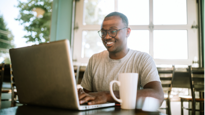A job candidate wearing glasses smiles while typing on a laptop, with a white mug next to it.