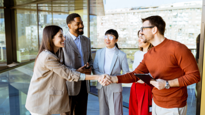 A group of five professionals stand in a modern office building with large glass windows. In the foreground, two colleagues smile and shake hands, while three others look on and engage in conversation. The scene conveys networking, collaboration, and building professional relationships in a workplace setting.