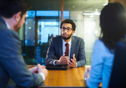 A job candidate in a suit and tie with glasses is asking questions to two interviewers across a table in an office setting.
