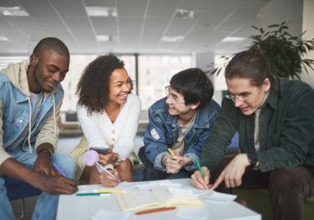 Four young adults sitting together at a table in a bright, modern office space, smiling and collaborating on a creative project with notebooks, papers, and colored pens