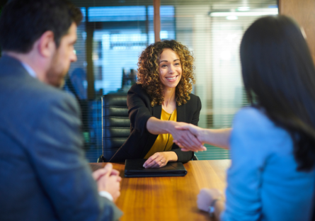 A job candidate sits across from two hiring team members. The candite is smiling and shaking hands with one of the hiring team members.
