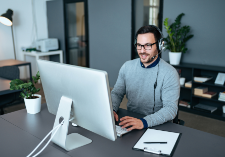 An IT helpdesk worker sits at his desk in from of a computer and talks into the headset he is wearing. There is a clipboard and pen on the desk to his left.