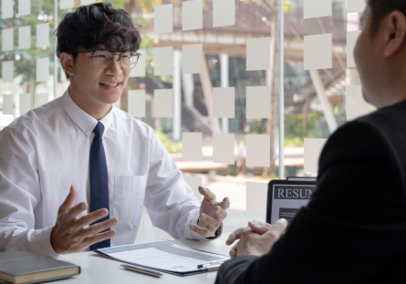 Man in a white shirt, dark tie, and glasses tells a story in an interview and uses his hands for emphasis. The interviewer, who wears a dark suit and is facing the candidate, listens with his hand folded in from of him.