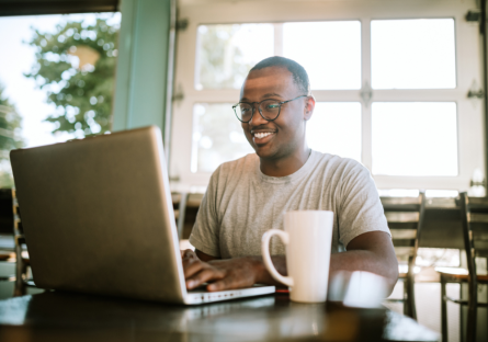 A job candidate wearing glasses smiles while typing on a laptop, with a white mug next to it.