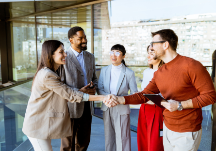 A group of five professionals stand in a modern office building with large glass windows. In the foreground, two colleagues smile and shake hands, while three others look on and engage in conversation. The scene conveys networking, collaboration, and building professional relationships in a workplace setting.