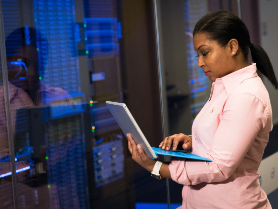 A software engineer is standing in front of a server studying her laptop that is in her hands. 