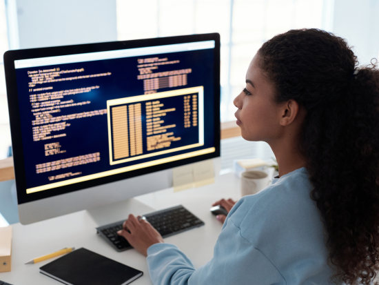 An IT worker sits in front of her computer that shows rows of code on the screen.