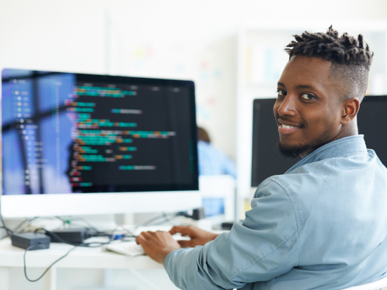A smiling IT worker sits in front of his computer that shows rows of code on the screen.