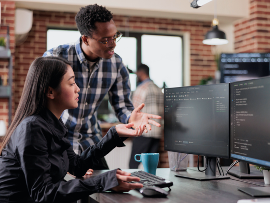 Two network technicians discuss the code that is on the two computer monitors in front of them.
