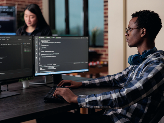A man who works in cybersecurity sits in front of two computer monitors, typing.