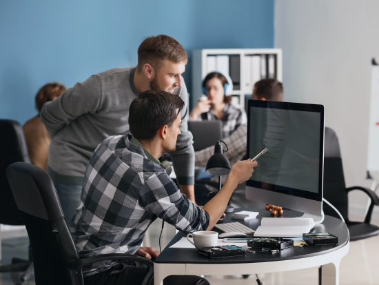 An IT worker leans over a colleague's computer, helping resolve his tech issues.