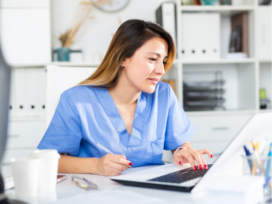 A medical administrative professional wearing blue scrubs works at a desk, typing on a laptop while reviewing paperwork in a bright, organized office setting.
