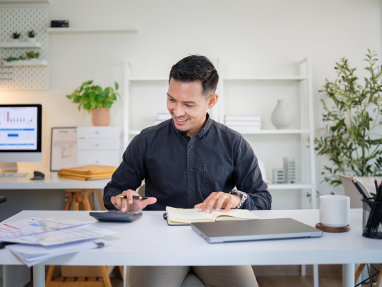 Person working at a desk reviewing a notebook and using a calculator, with a laptop and financial documents nearby, representing bookkeeping or accounting work in a modern office setting.