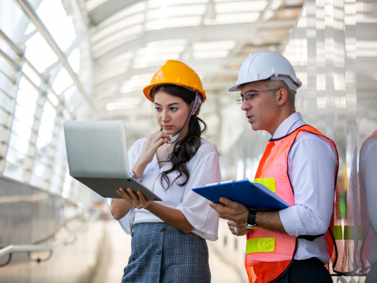 Two manufacturing technologists in hard hats stare at a laptop and clipboard.