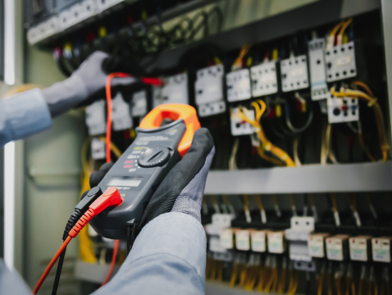 Electrical technician using a clamp meter to test voltage inside an electrical control panel.