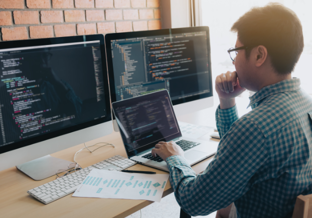 Male working on a laptop with two desk monitors comparing data.