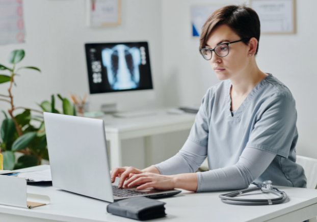 Female medical administrative assistant in scrubs working on a laptop in an office setting.