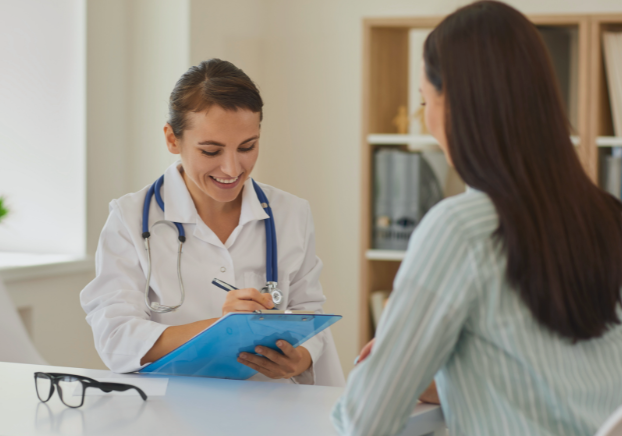 Female healthcare professional in a white coat with a stethoscope around their neck writing on a paper attached to a clipboard and discussing information with a female patient.